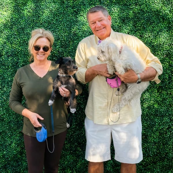 A smiling couple holding their two small dogs in front of a green hedge wall.