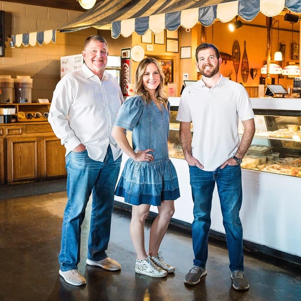 Three smiling people, two men and one woman, stand in a restaurant with a food display case.