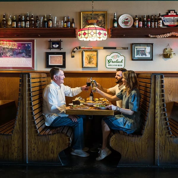 Three people toast drinks in a cozy restaurant booth filled with food and vintage decor.