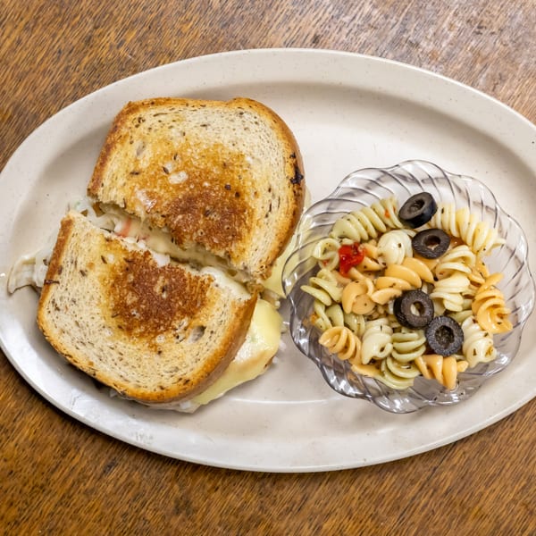 A grilled deli sandwich with melted cheese and a side of pasta salad with black olives.