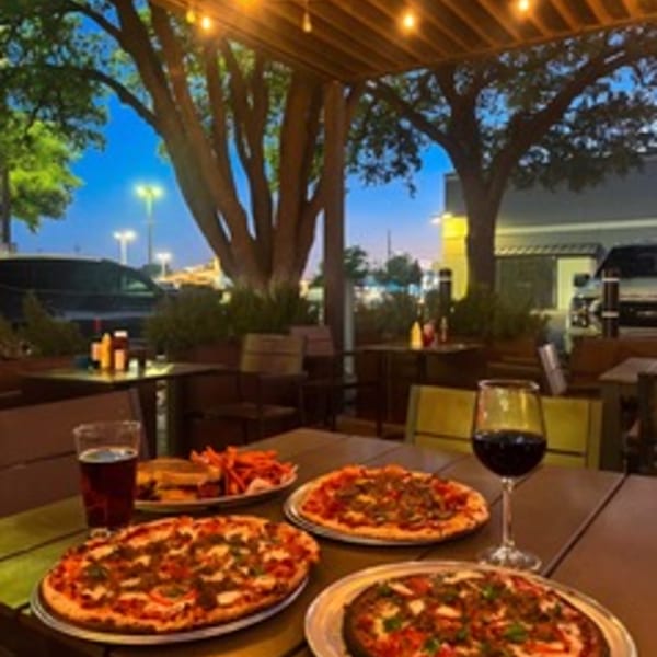 Outdoor patio at dusk with string lights, three pizzas, a burger with fries, and drinks on a table.