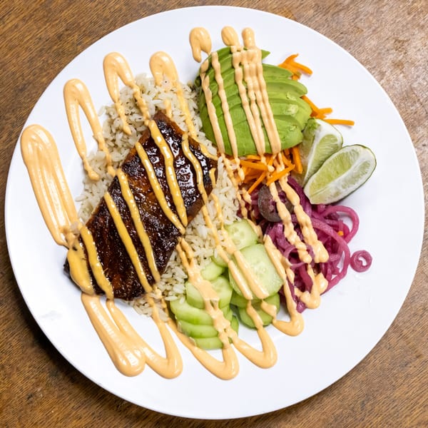 A deli bowl with glazed meat, rice, avocado, pickled onions, cucumbers, and lime, drizzled with sauce.