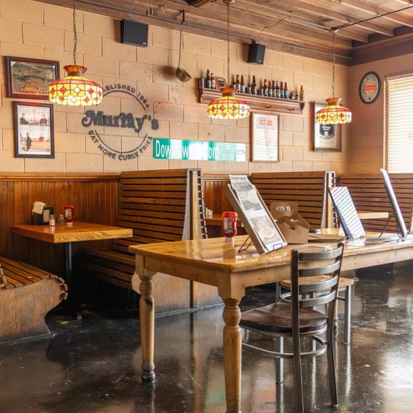 Interior of Murray's Deli with wooden tables, booths, and stained-glass lamps under a brick wall.