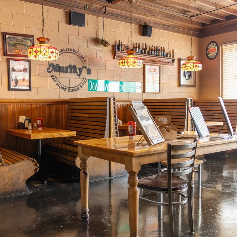 Interior of Murray's Deli with wooden tables, booths, and stained-glass lamps under a brick wall.